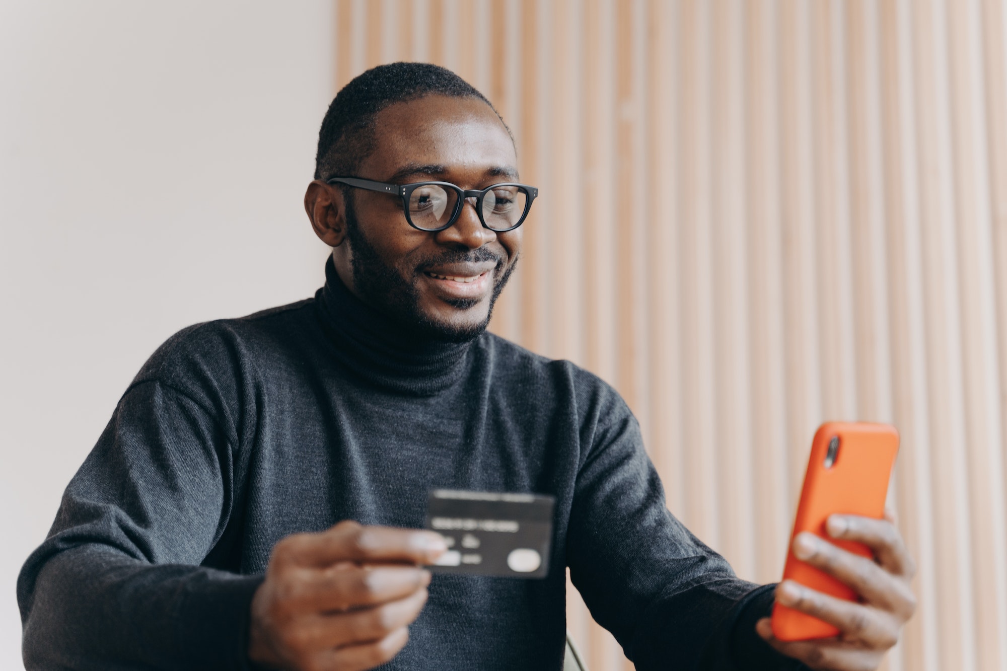 Young happy african american male entrepreneur with credit card using e-banking app on smartphone